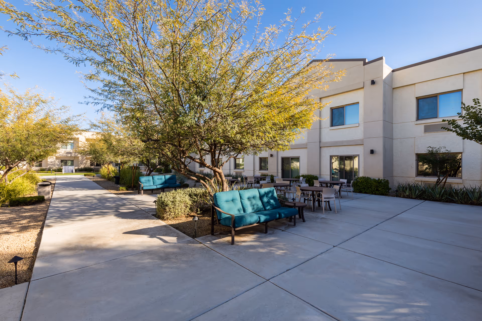 Sunlit courtyard with teal outdoor sofas, tables, trees, and a two-story beige building in the background.