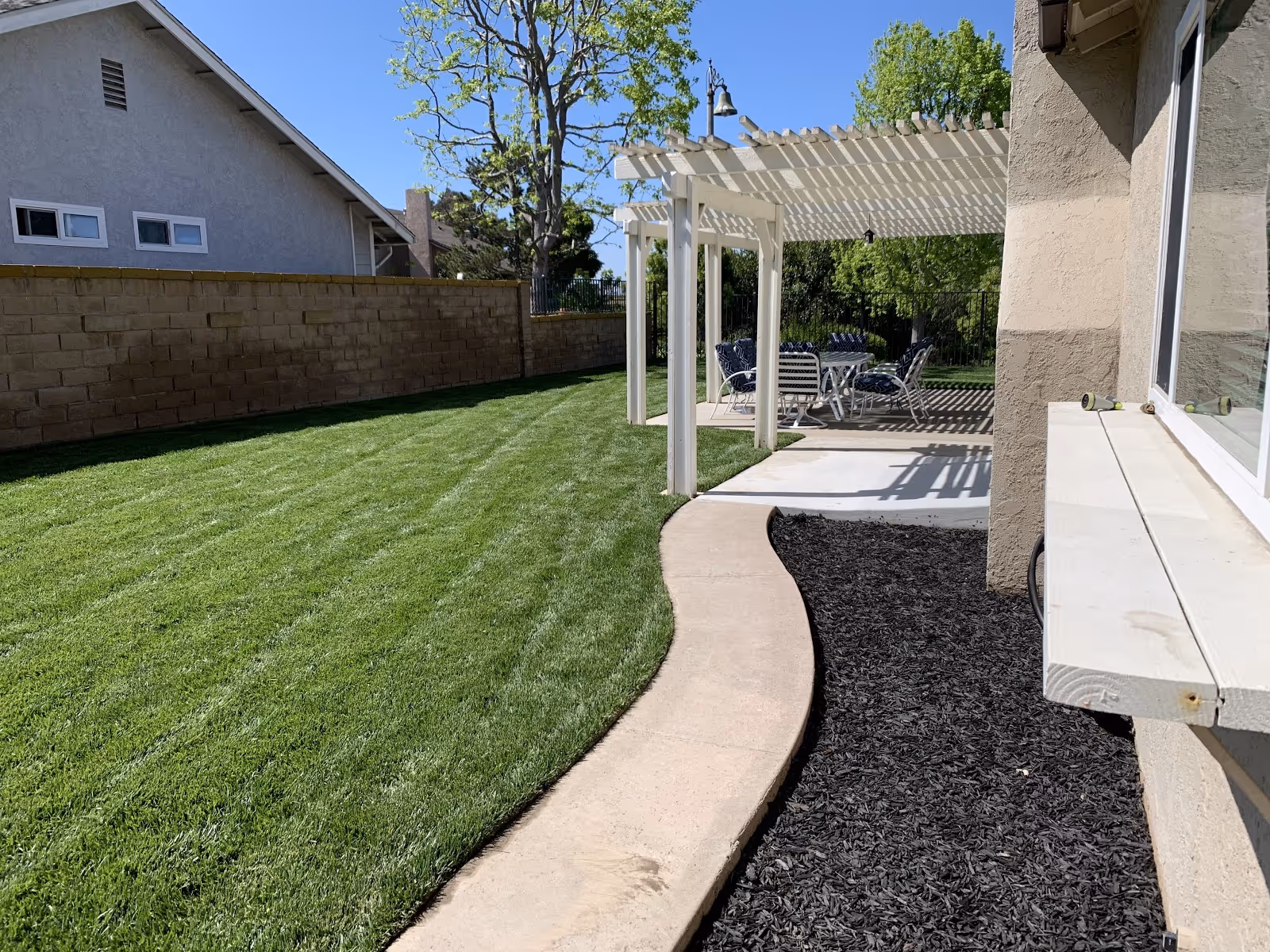 A backyard area with a well-maintained green lawn, a curved concrete pathway, and a white pergola providing shade over a patio set with chairs and a table. There is a beige building wall on the right side and a brick wall enclosing the yard on the left. Trees and a clear blue sky are visible in the background.