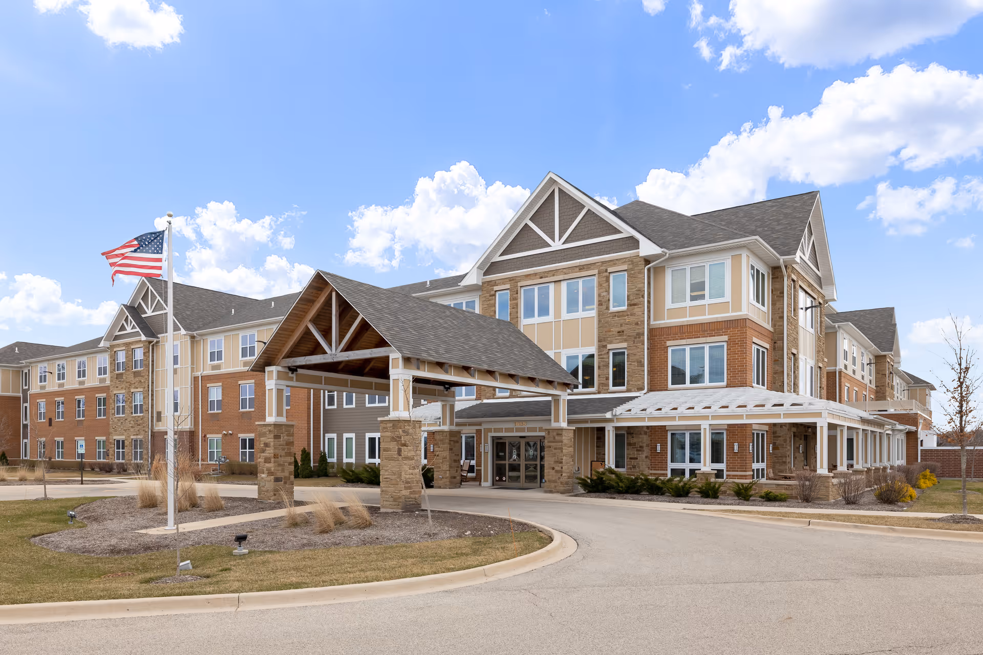 Exterior view of a multi-story senior living facility building with a covered entrance, an American flag on a flagpole, and a clear blue sky with some clouds.