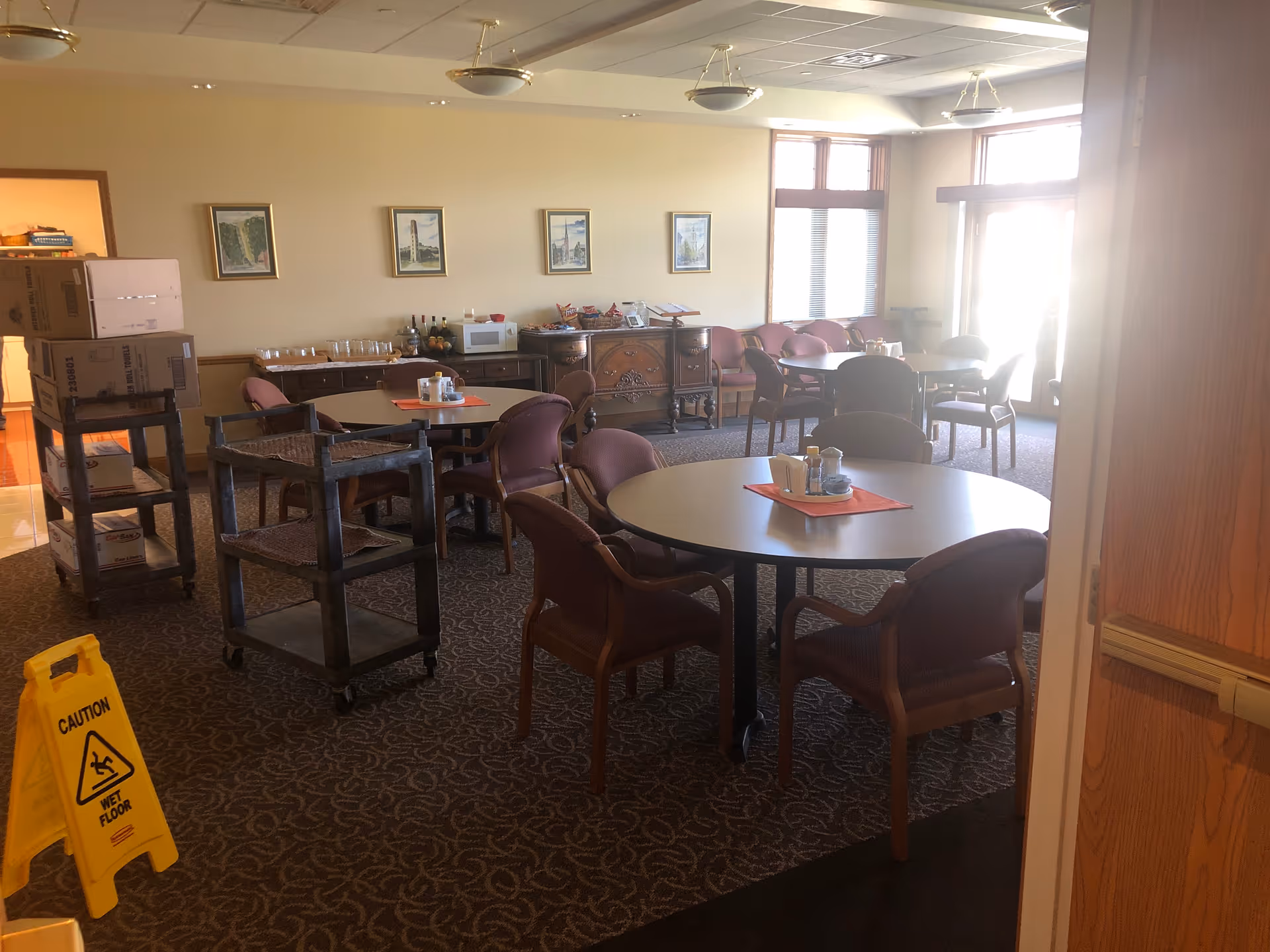 A senior living dining room with round tables, chairs, service carts, and a yellow caution sign.