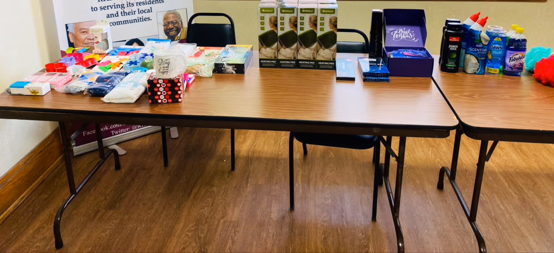 Long folding tables in a room display assorted household supplies and toiletries.