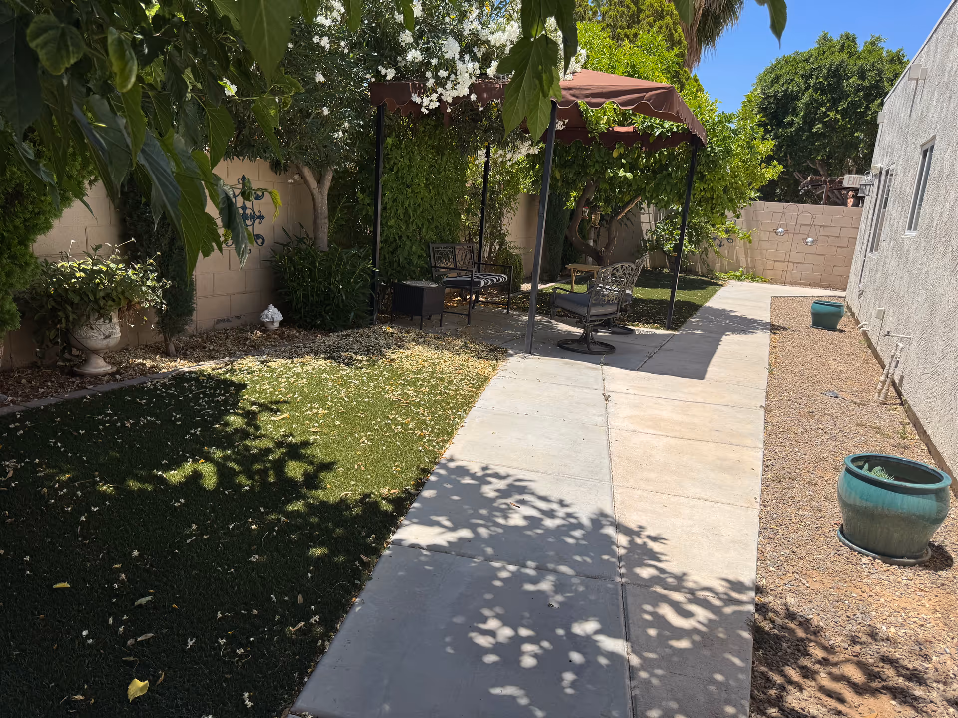 Outdoor garden area with a concrete pathway leading to a shaded seating area with metal chairs and a small table. The garden has green plants, trees, and potted plants along the sides, with a beige wall enclosing the space. The sky is clear and blue.