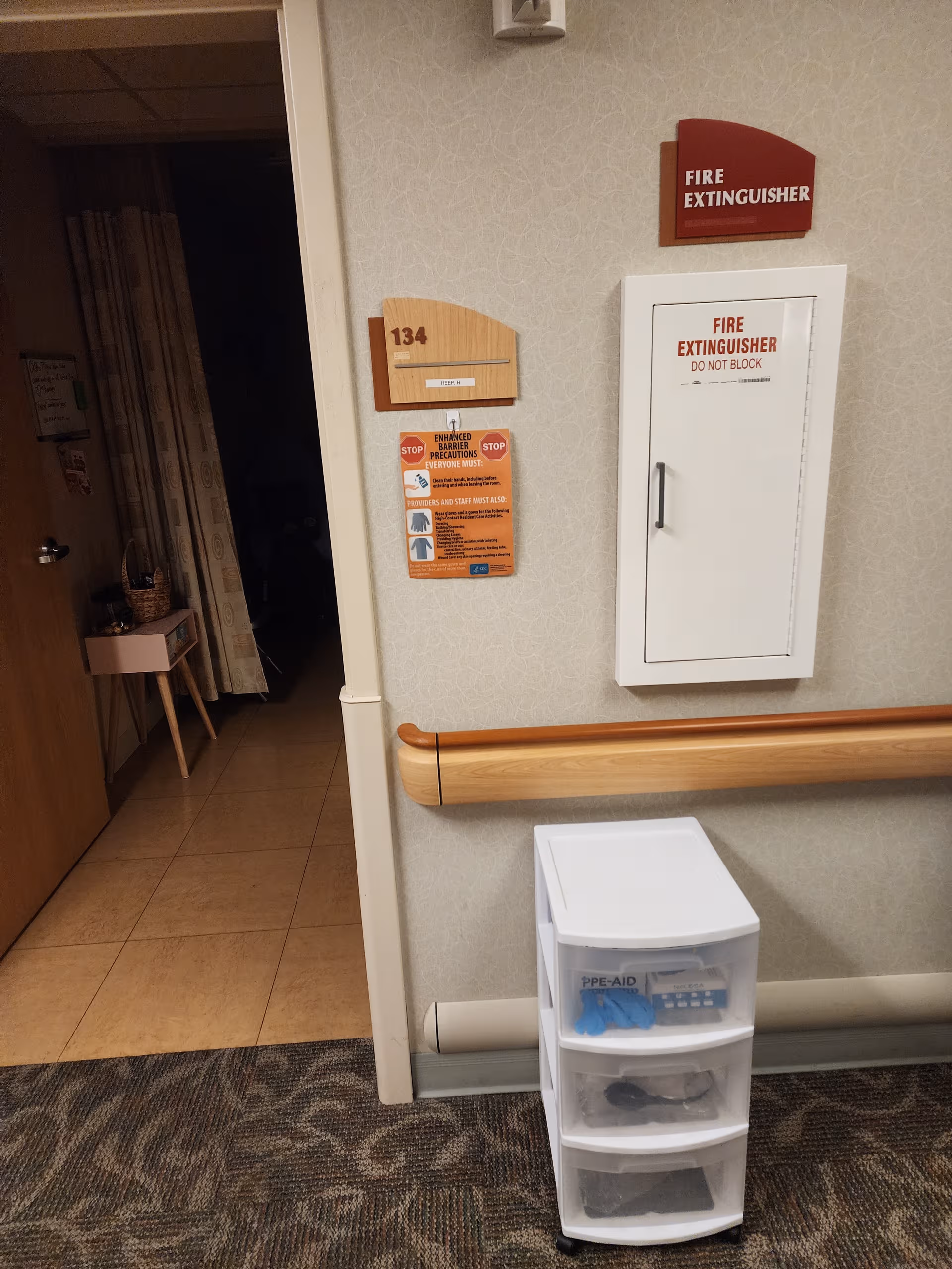 Interior hallway with wall-mounted fire extinguisher cabinet, wooden handrail, and PPE supply drawer