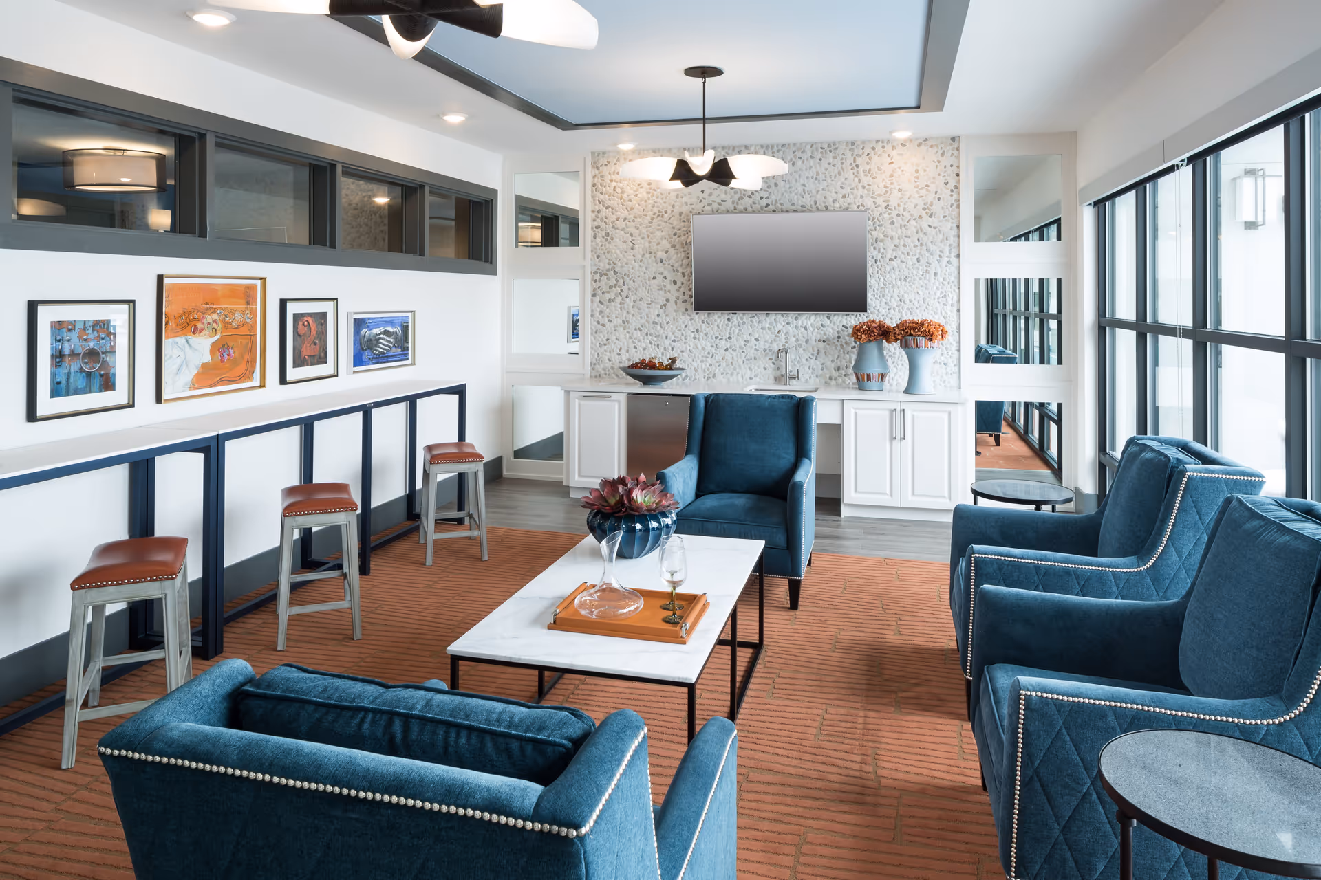 Bright communal lounge with blue armchairs around a marble coffee table, bar stools along a counter, and large windows.