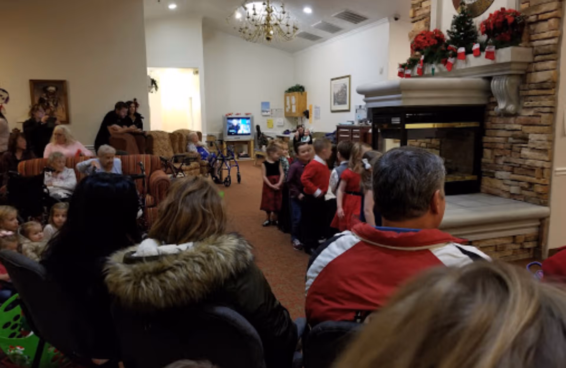 A group of children dressed in festive clothing standing in a line near a stone fireplace decorated with Christmas decorations, while adults and elderly people sit on chairs and couches watching them in a cozy living room setting.