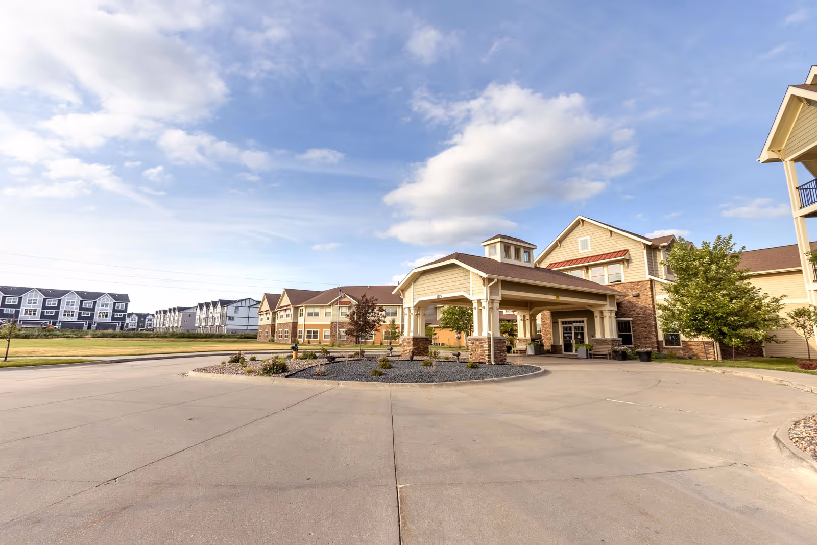 Front exterior of a senior living facility with a covered porte-cochere and adjacent buildings under a blue sky.