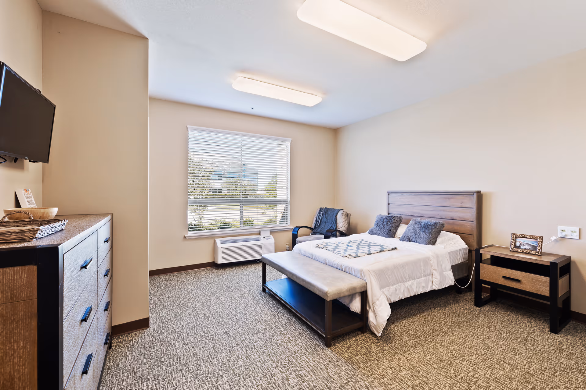 A furnished bedroom in a senior living facility with a bed, dresser, bench, and window.