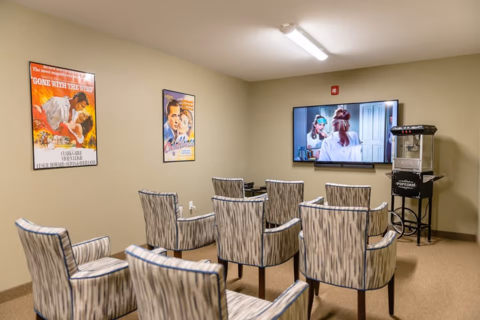 Small media room with rows of patterned chairs facing a wall-mounted TV and a popcorn machine, with movie posters on the wall.