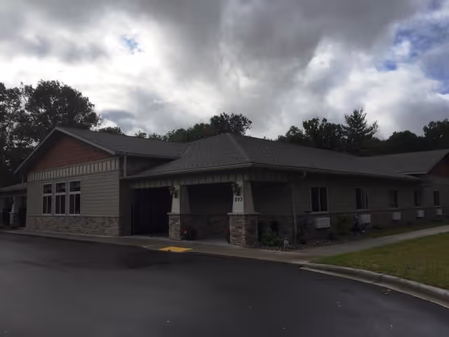 Exterior view of a single-story senior living facility building with a covered entrance, stone and siding exterior, surrounded by trees under a cloudy sky.