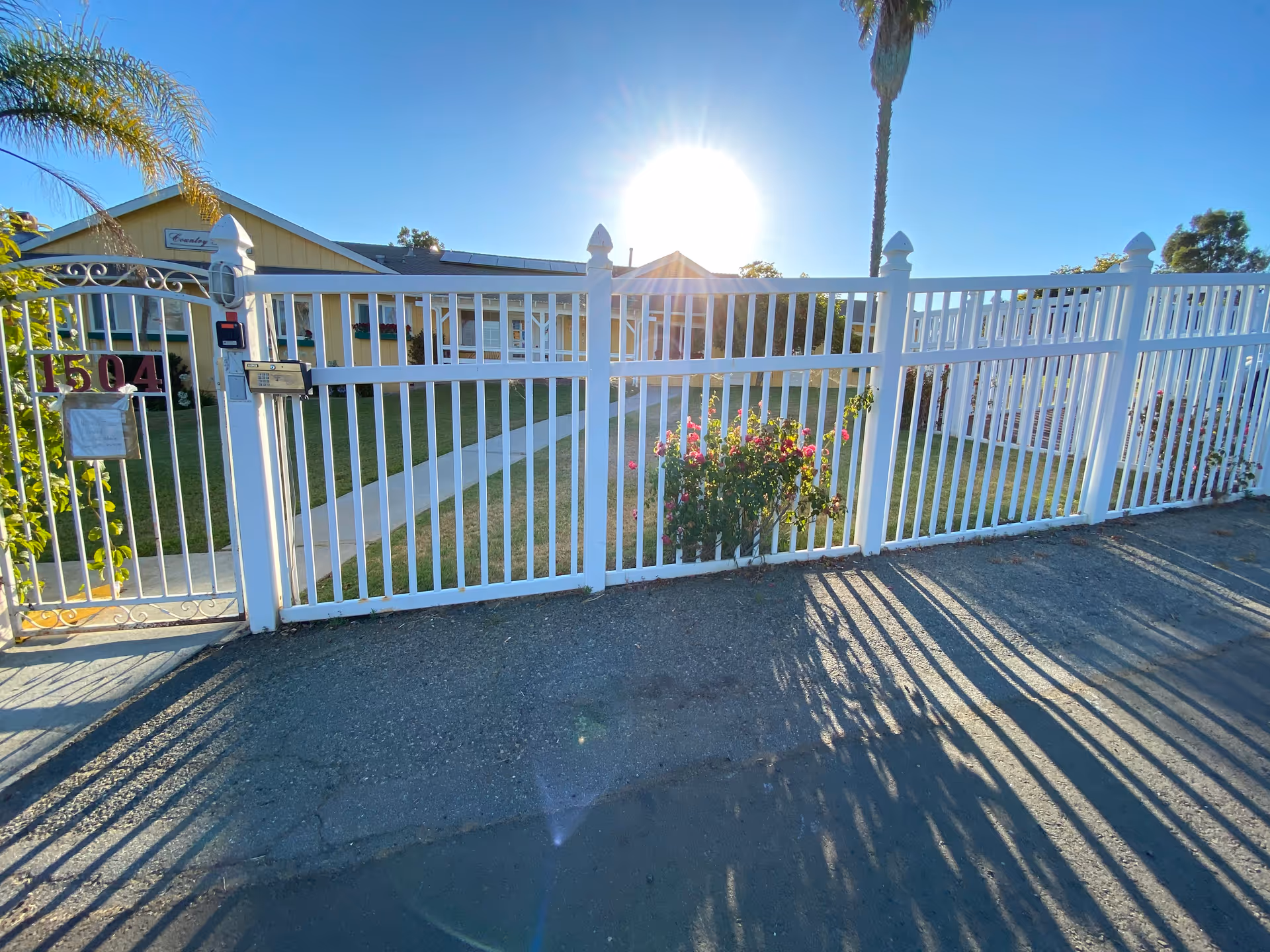 White metal gate and fence in front of a sunny senior facility yard.