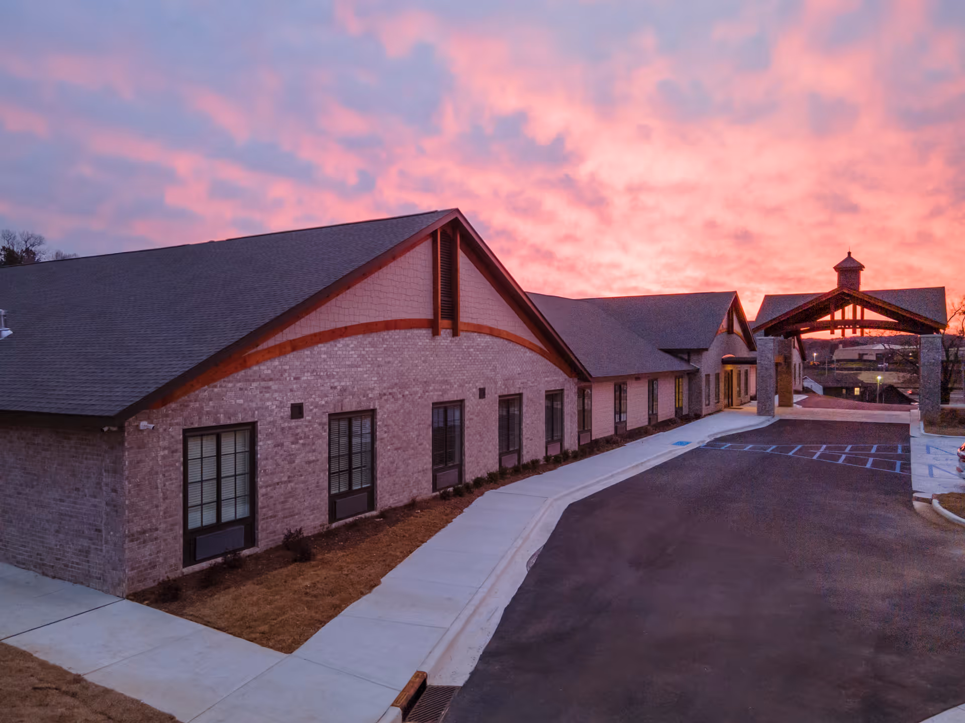 Sunset sky above a brick senior living facility exterior with a curved sidewalk and driveway.