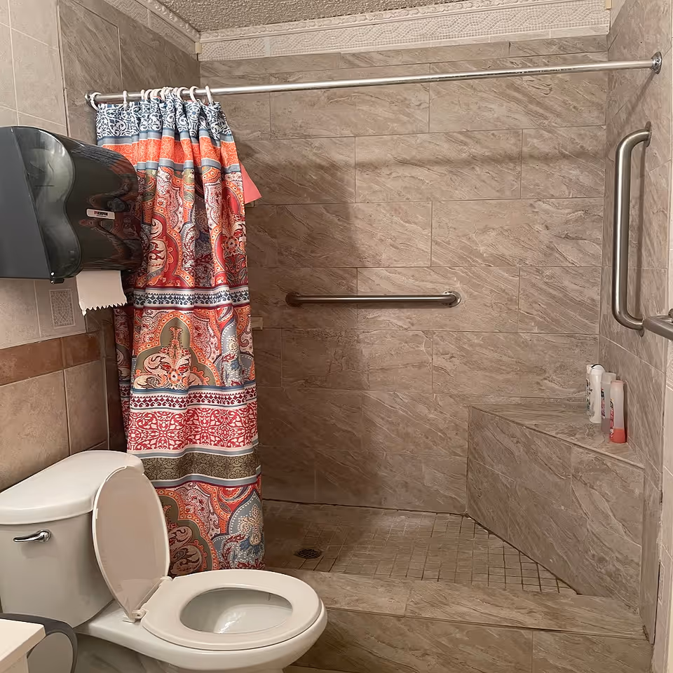 A tiled bathroom with a shower, colorful curtain, grab bars, and toilet in a senior facility.