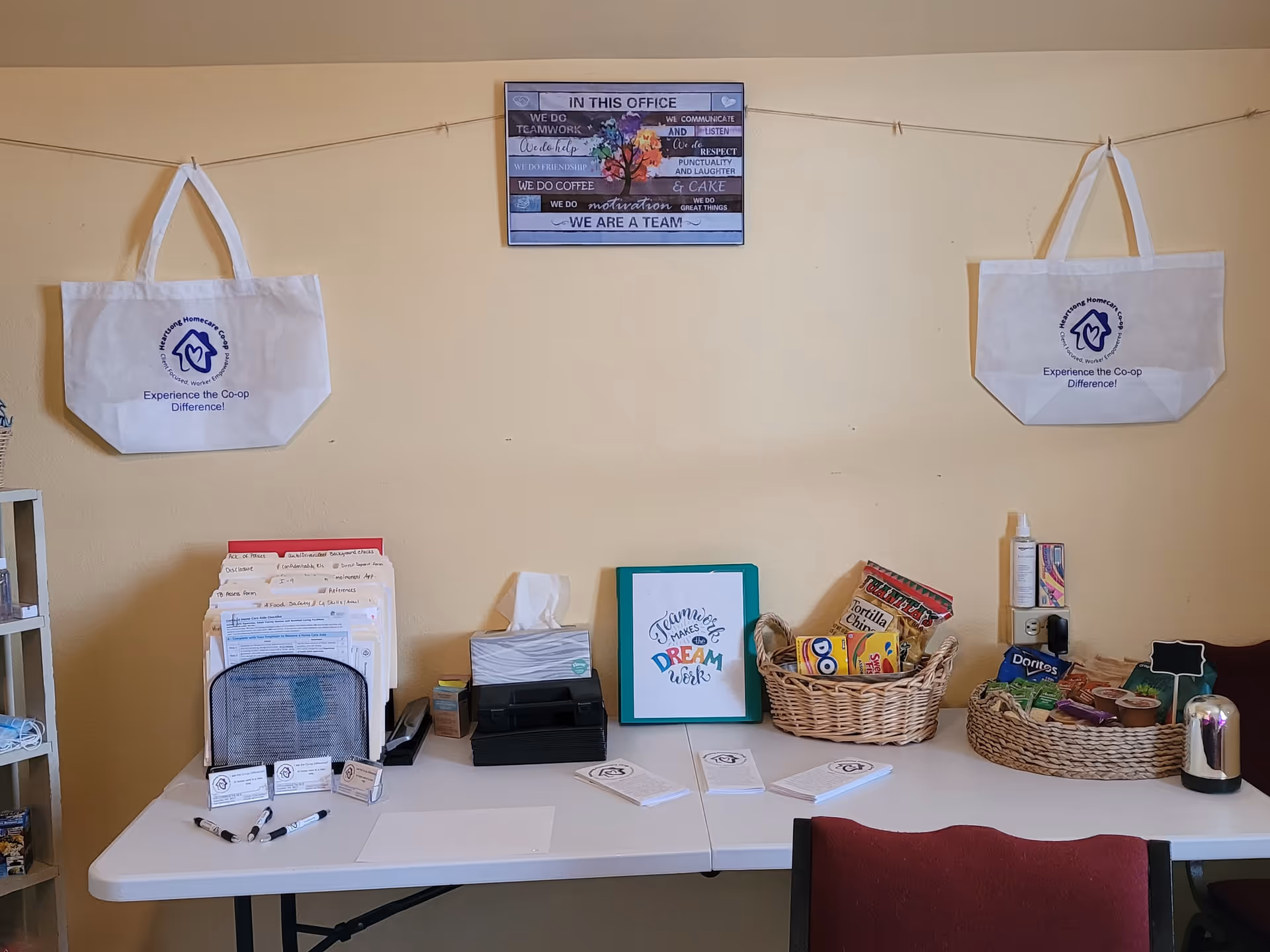 Office desk with baskets, papers, and white tote bags hung on a pale yellow wall.