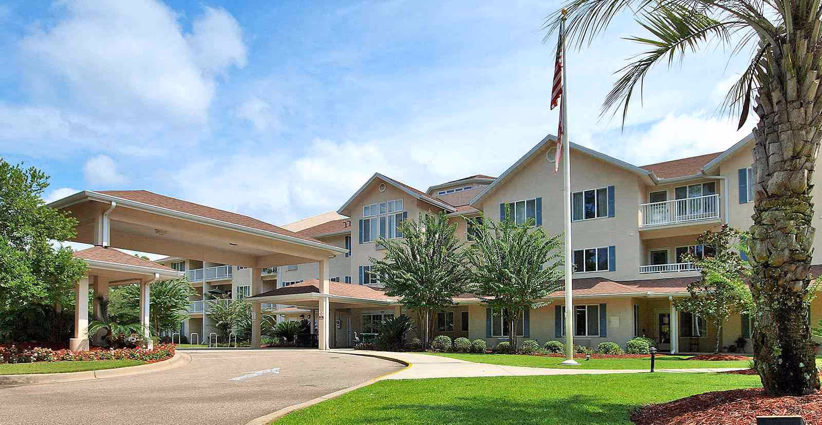 Front exterior of a senior living building with a covered porte-cochere, flagpole, palm tree, and landscaped lawn.