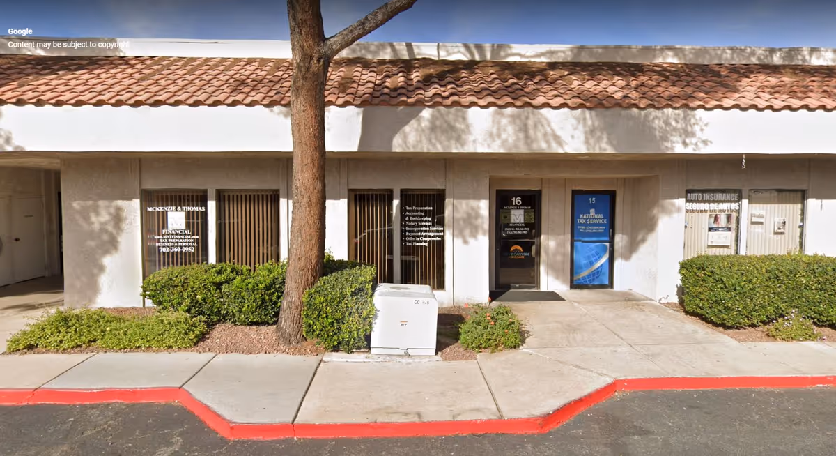 Exterior front of a strip-mall building with storefronts, shrubs, and a central tree.