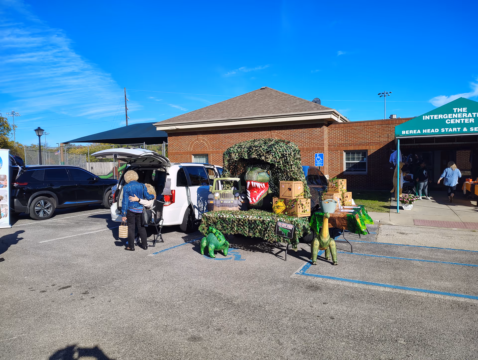 People near a vehicle in a parking lot beside a brick building with a dinosaur display.