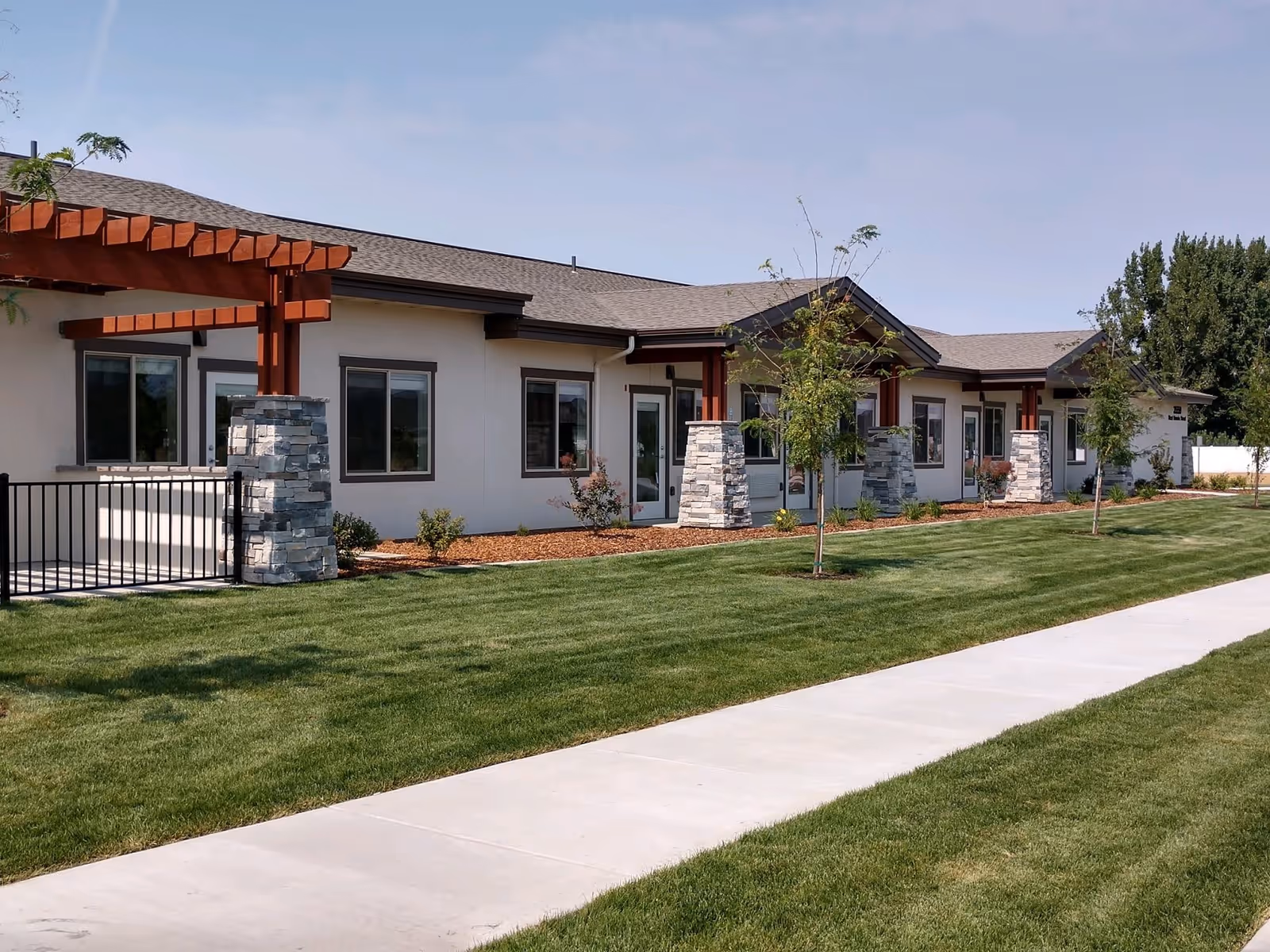Single-story senior living building exterior with covered entries supported by stone columns, a manicured lawn, and a concrete sidewalk.