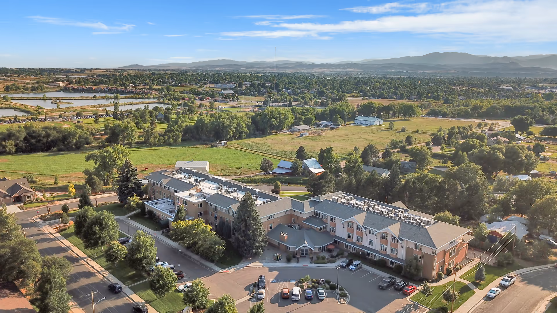 Aerial view of Hillcrest Of Loveland senior living facility surrounded by green fields, trees, and residential areas with mountains in the background under a partly cloudy sky.