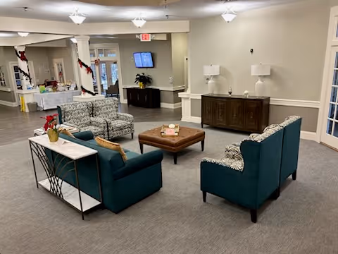 A senior living facility common area with teal and patterned armchairs arranged around a square wooden coffee table. There is a console table with a small plant and decorative items, and a sideboard with two lamps against the wall. The room has neutral-colored walls and carpeted flooring, with a hallway and entrance visible in the background.