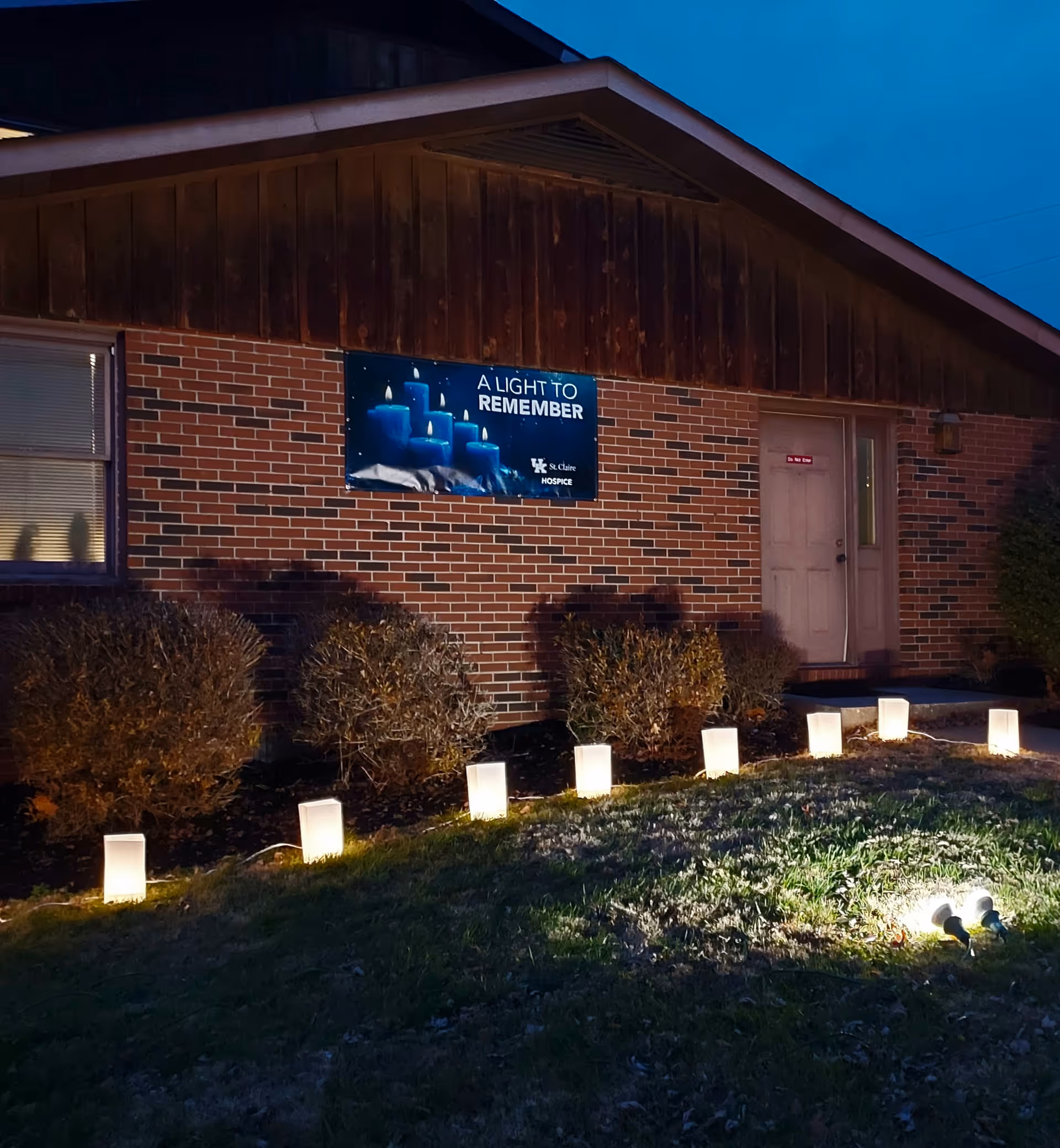 Exterior view of UK St. Claire Hospice, brick building at dusk with banner and path lights.