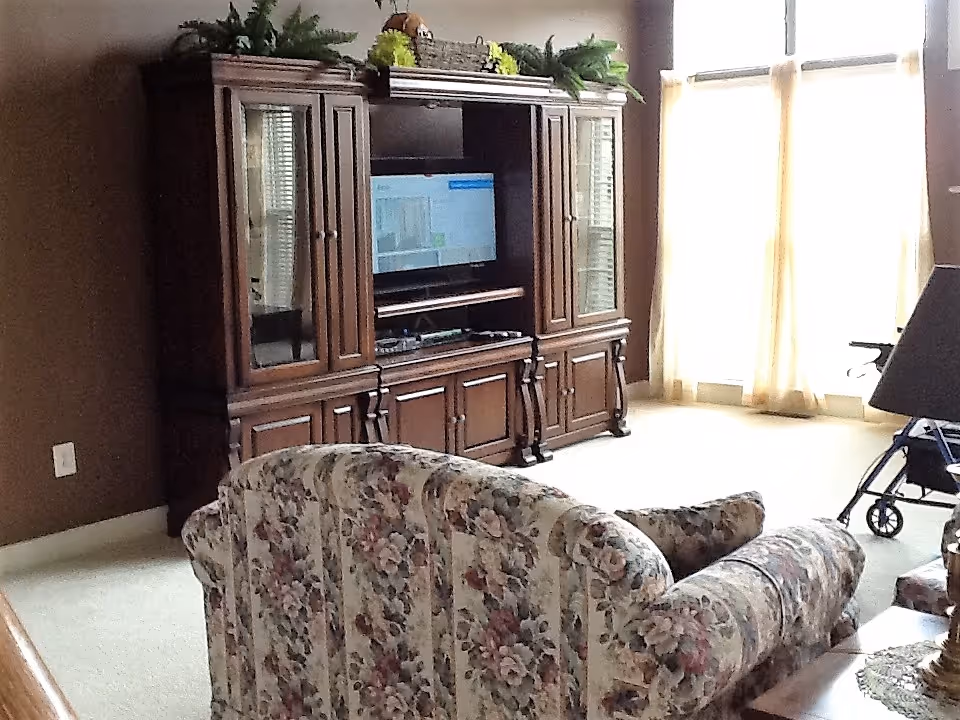 Living room with a floral patterned sofa facing a large wooden entertainment center with a TV. The room has beige carpet, brown walls, and large windows with sheer curtains letting in natural light. A walker is visible near the window.
