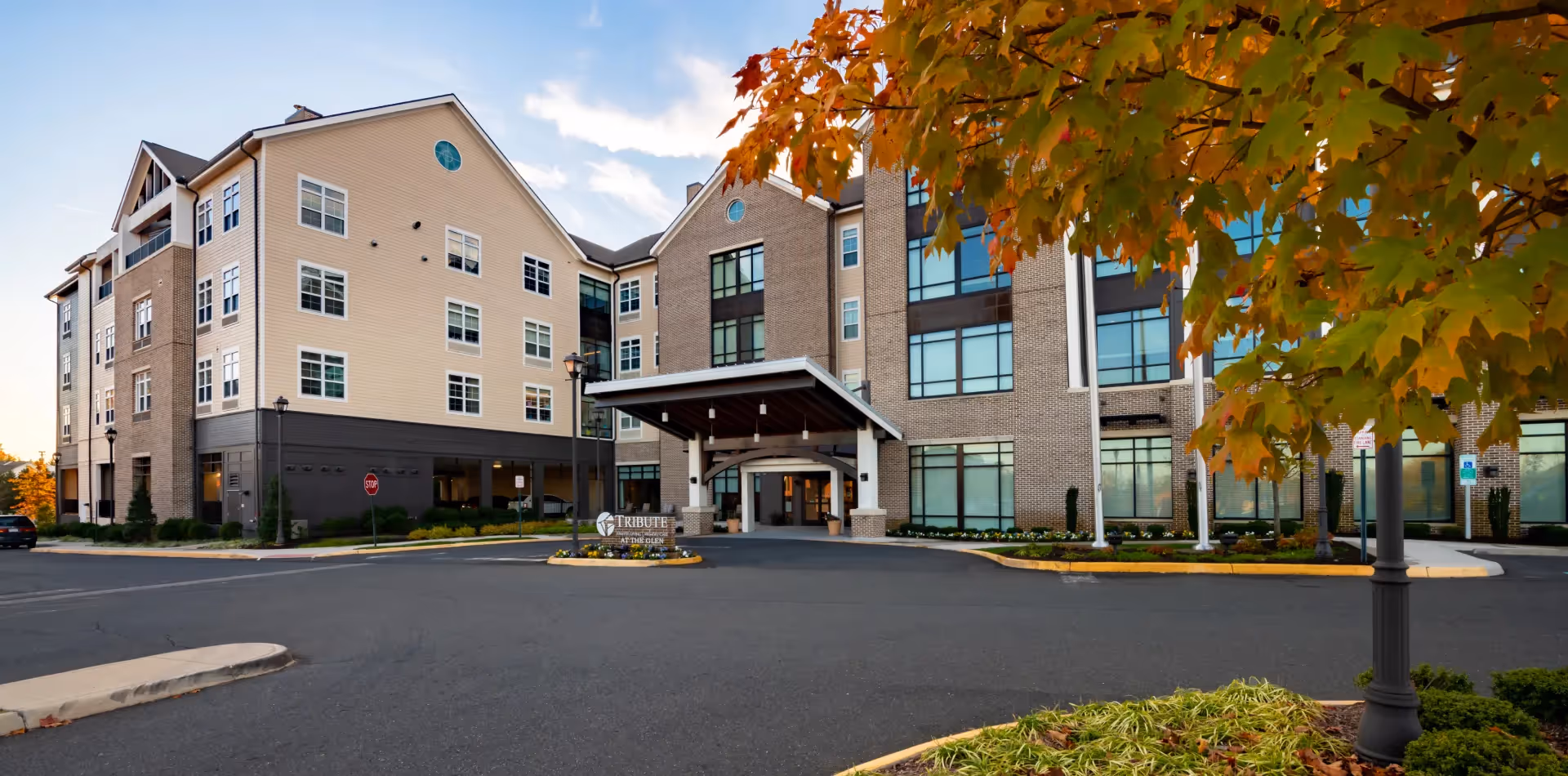 Front exterior of the Tribute at The Glen senior living building with a covered entrance, circular driveway, and autumn foliage in the foreground.
