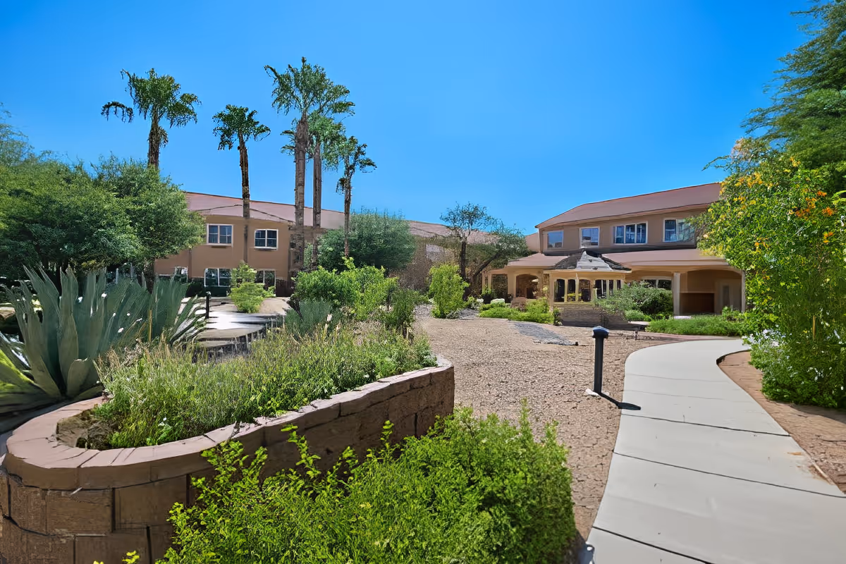 Landscaped courtyard with a curved pathway leading to the front of a two-story assisted living building surrounded by palm trees and desert plants.