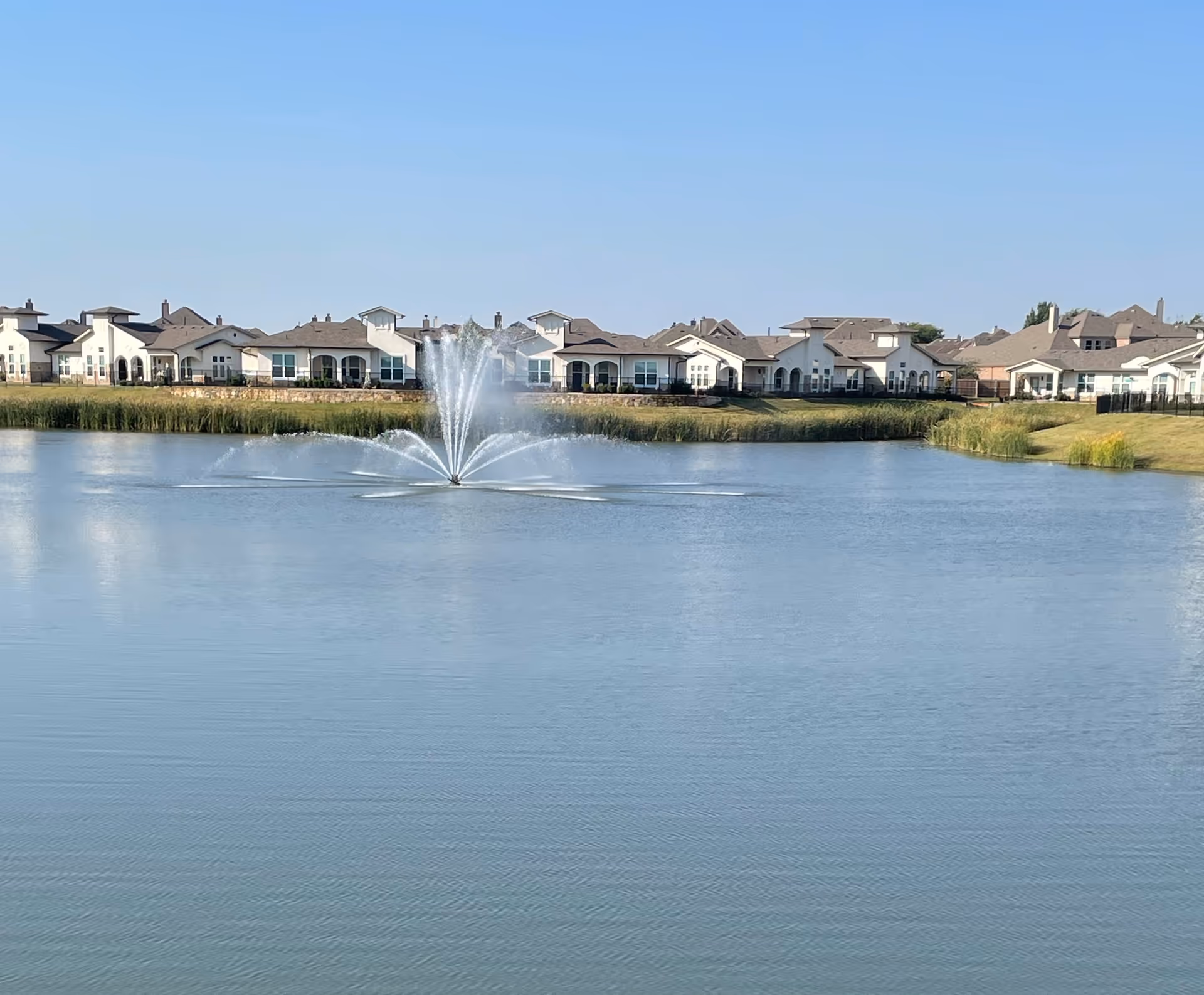 Water fountain in a lake with a row of white senior living buildings in the background.