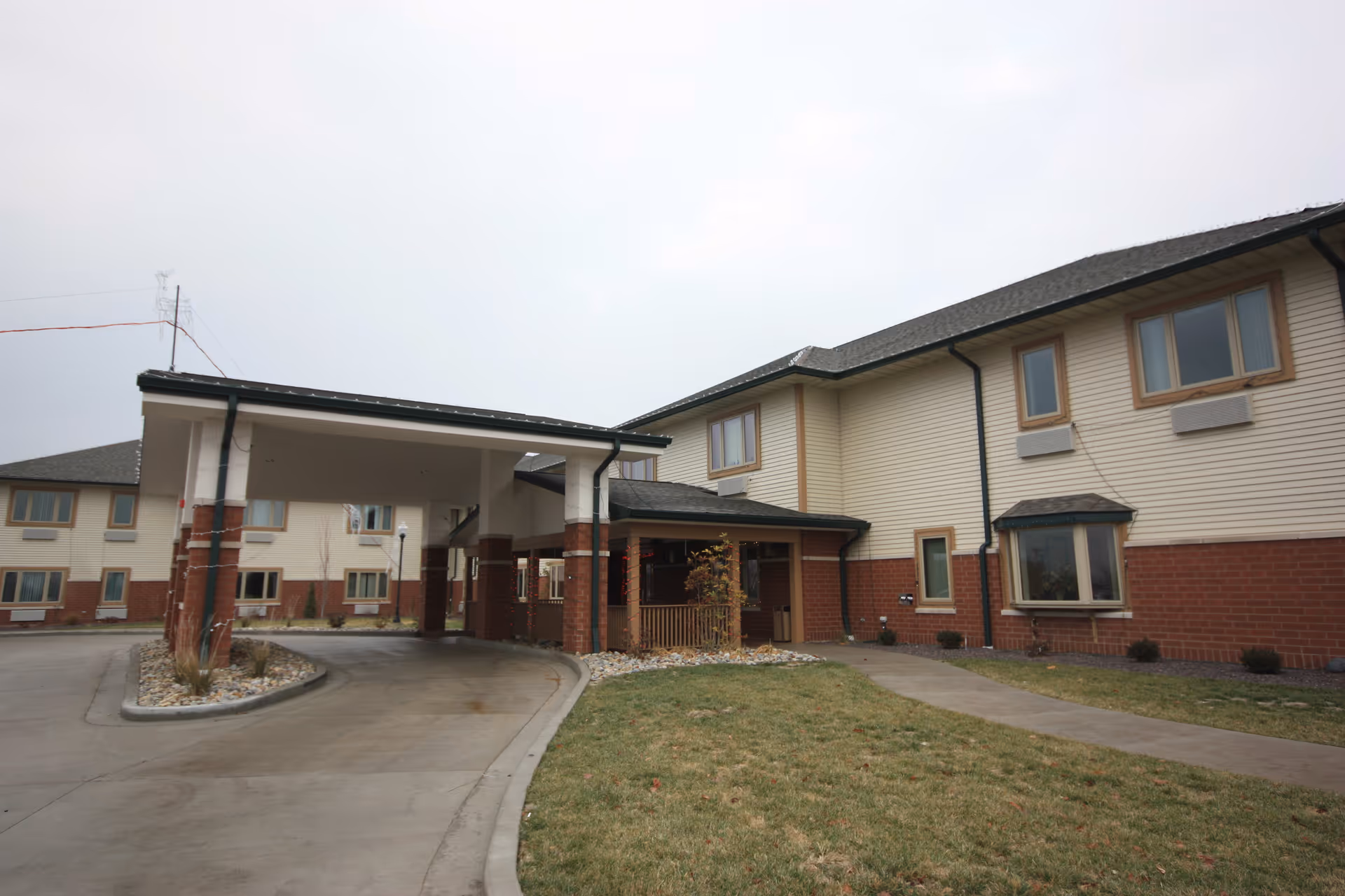 Exterior view of a two-story senior living facility building with beige siding and red brick accents. There is a covered driveway entrance with pillars and a curved concrete driveway. The surrounding area has some grass and small landscaped areas with rocks and plants.