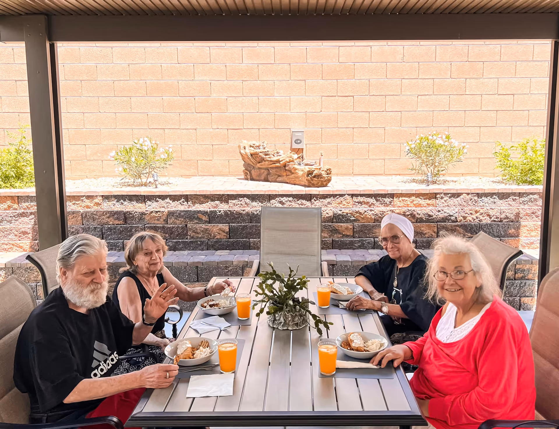 Four elderly people sitting around a rectangular outdoor table under a covered patio, enjoying a meal with plates of food and glasses of orange juice. The background shows a brick wall with some small plants and a decorative wooden sculpture.