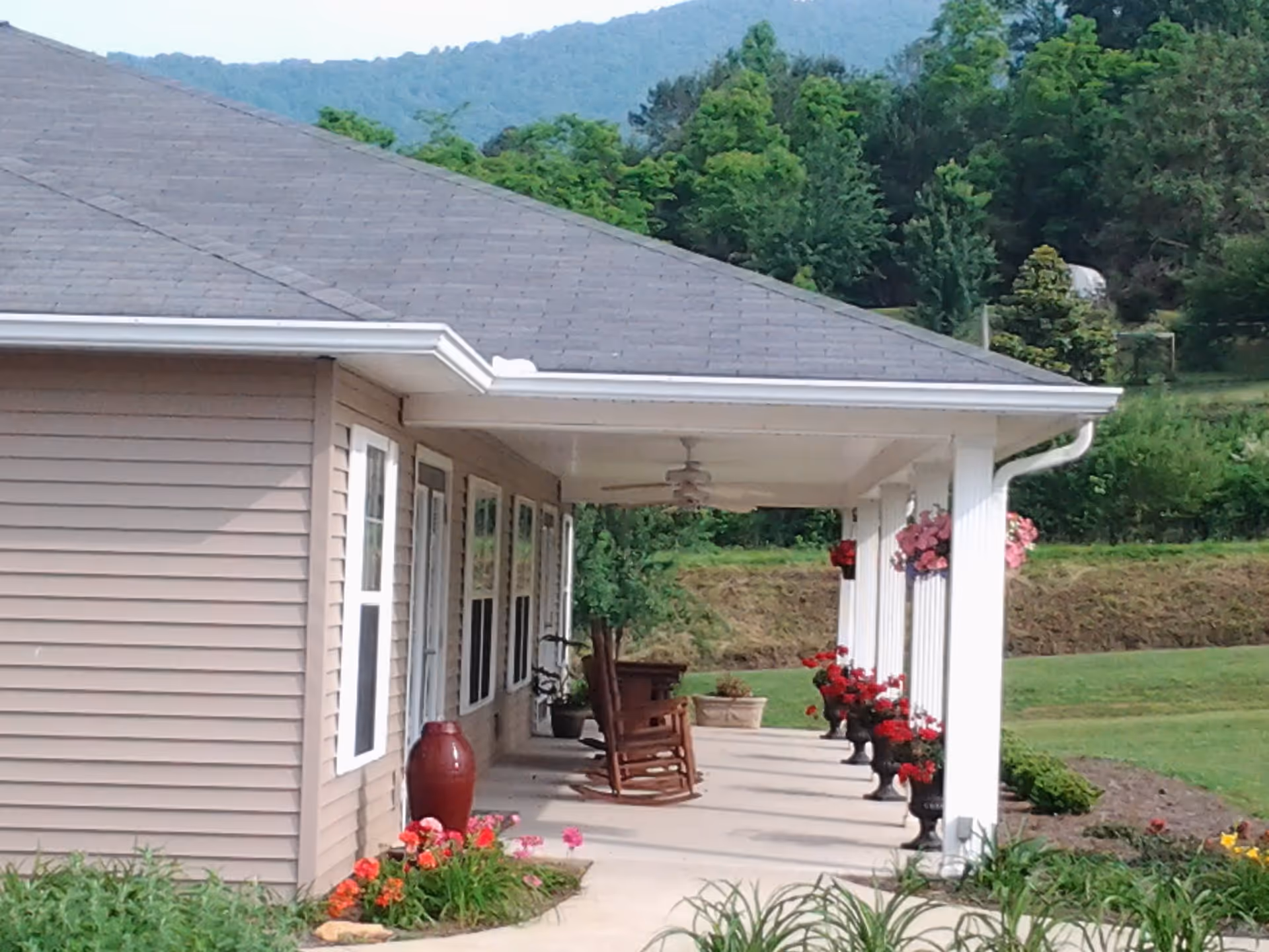 Covered front porch of a single-story care home with rocking chairs, potted flowers, and a lawn with hills in the background.