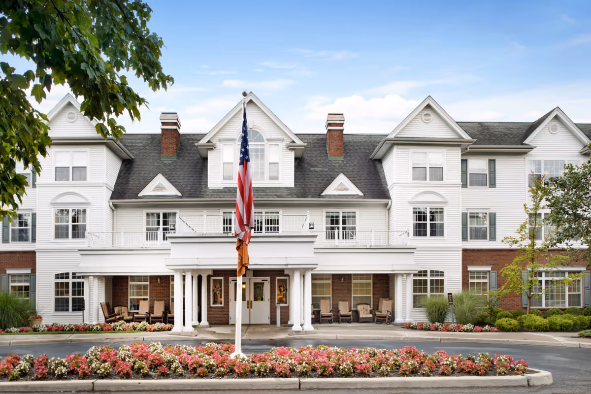 White multi-story senior living building with flagpole, porch seating, and flower beds in front.