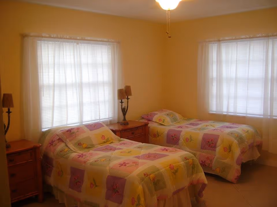 A bedroom with two twin beds covered in colorful floral-patterned quilts. Each bed has a wooden nightstand beside it with a lamp. The room has two large windows with sheer white curtains, and the walls are painted a soft yellow.