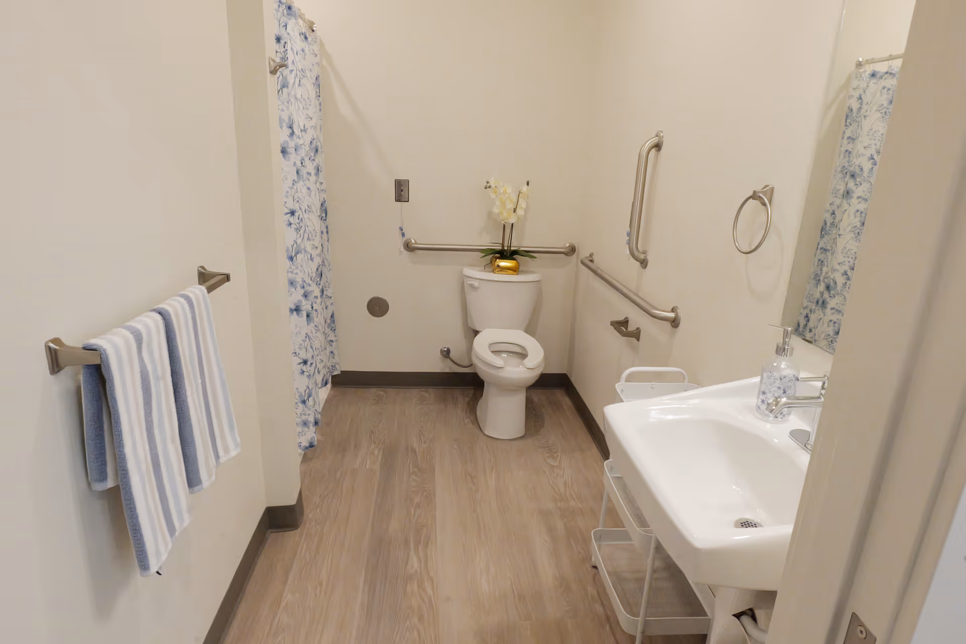 Accessible bathroom featuring toilet, sink, grab bars, and blue floral shower curtain.