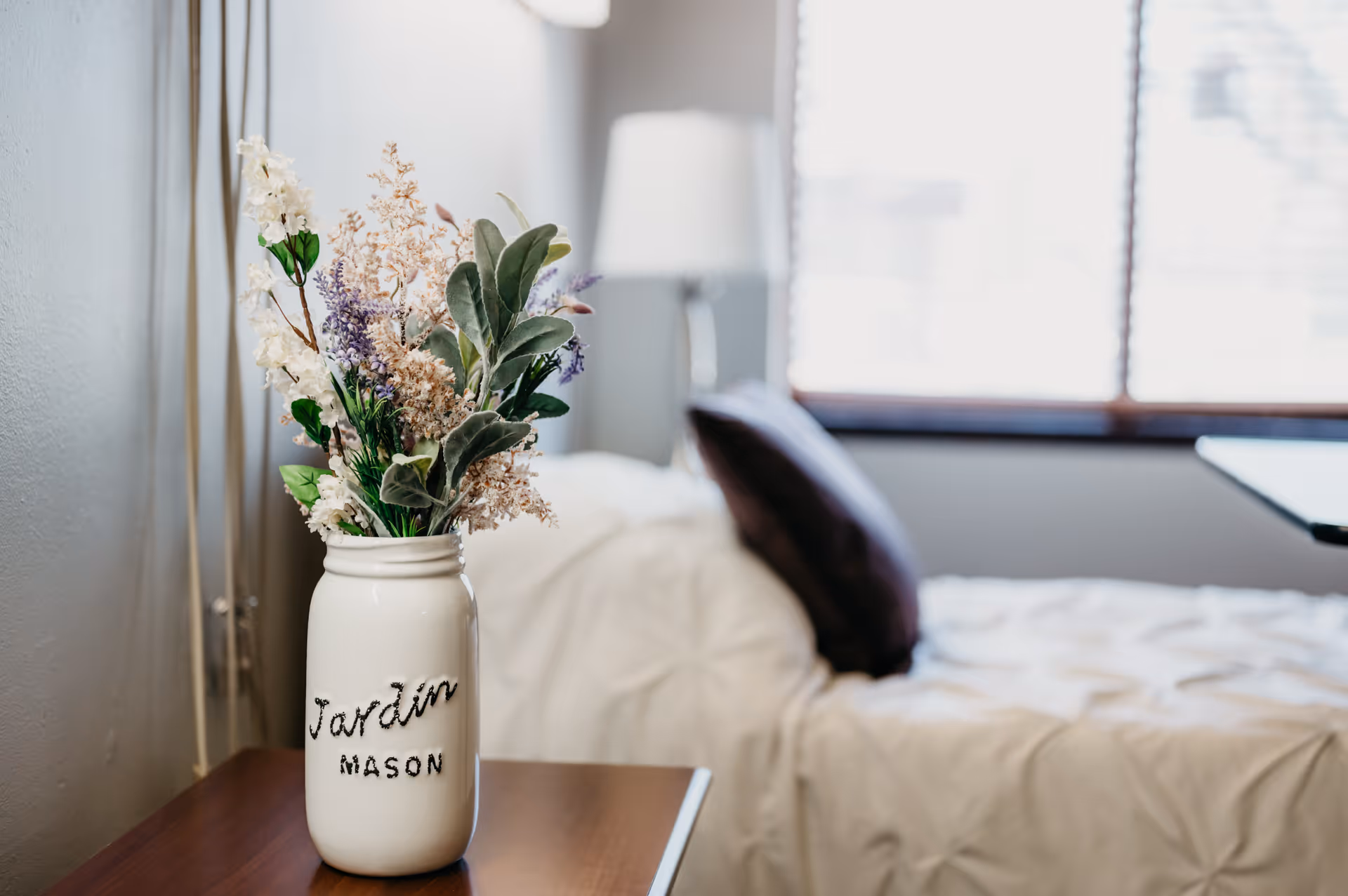 A vase with dried flowers sits on a wooden nightstand beside a neatly made bed.