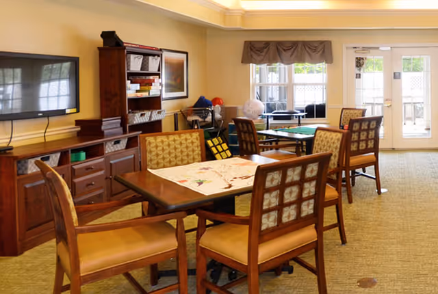 Well-lit common room with wooden tables and chairs, a TV and shelving stocked with games and supplies.