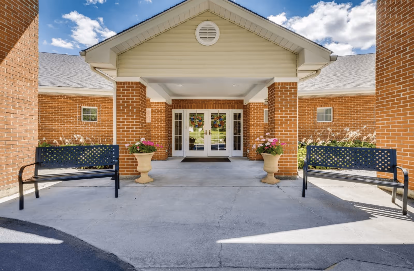Entrance to a brick building with a covered porch supported by two brick columns. There are two black metal benches on either side of the entrance and two large beige planters with pink and white flowers. The sky is blue with some clouds.