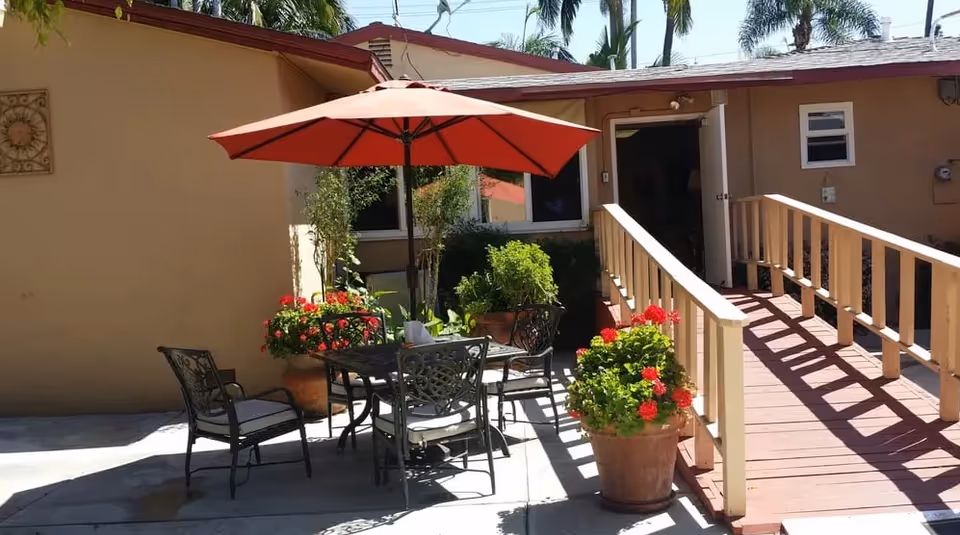 Outdoor patio area with a table and four chairs under a red umbrella, surrounded by potted plants with red flowers. There is a wooden ramp leading to a door of a beige building with a red roof.