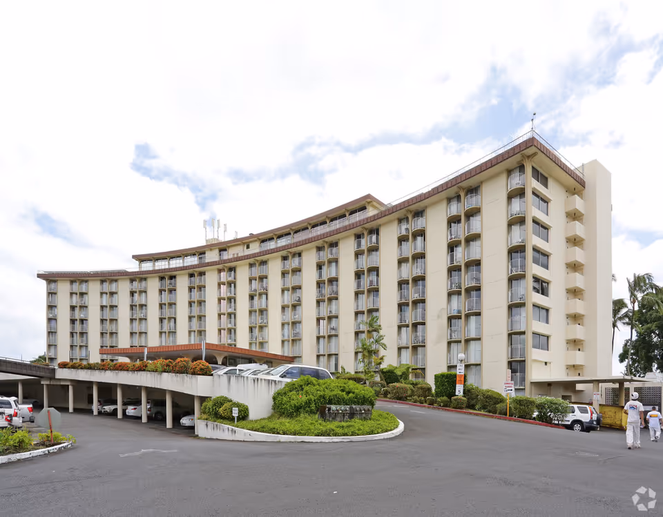 A large curved senior living facility building with many balconies and a parking lot in foreground.