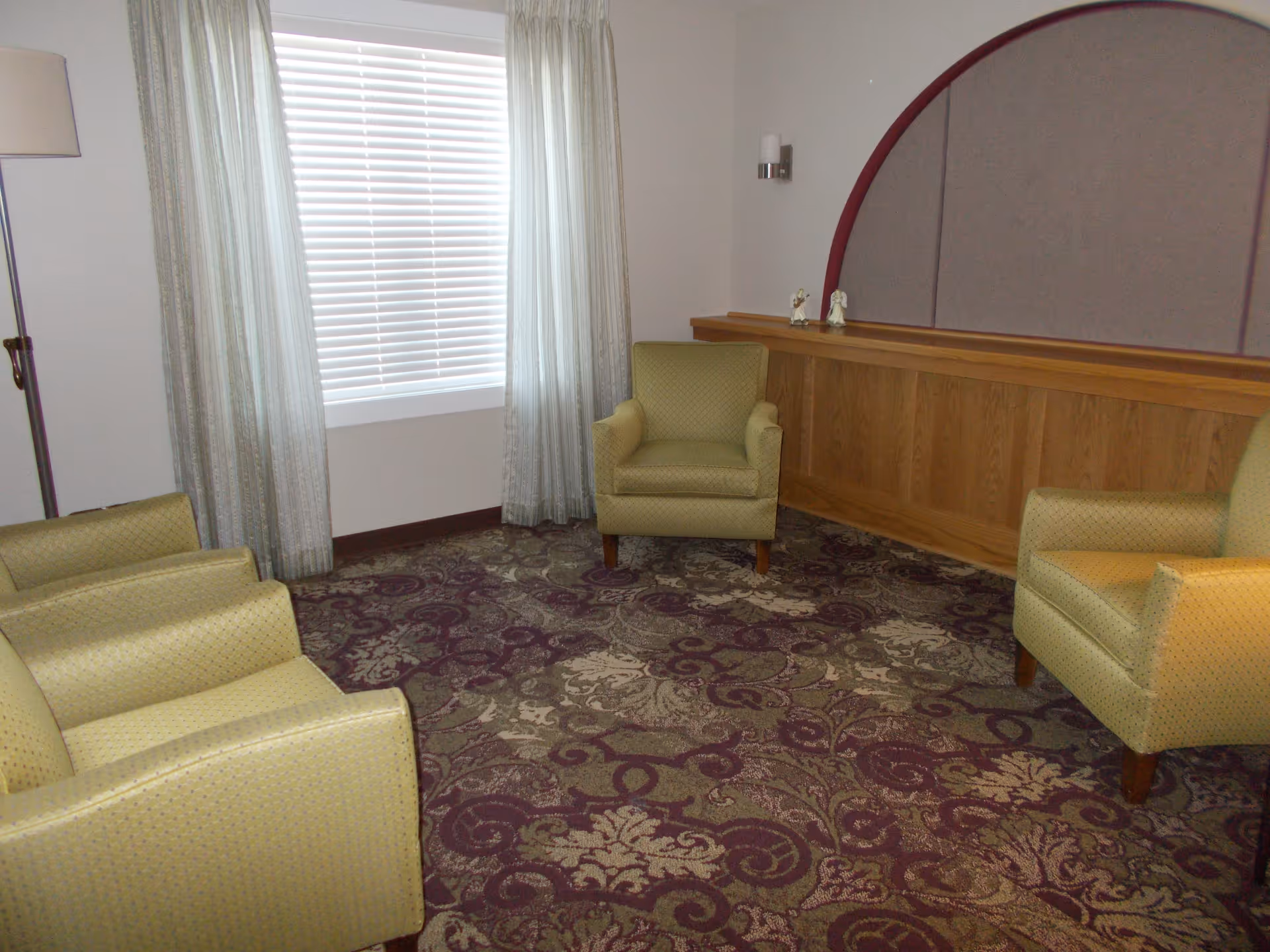 A small sitting area with four light green upholstered armchairs arranged around a patterned carpet with purple and beige floral designs. There is a window with white blinds and light curtains, a floor lamp, and a wooden panel with a decorative arch on the wall.