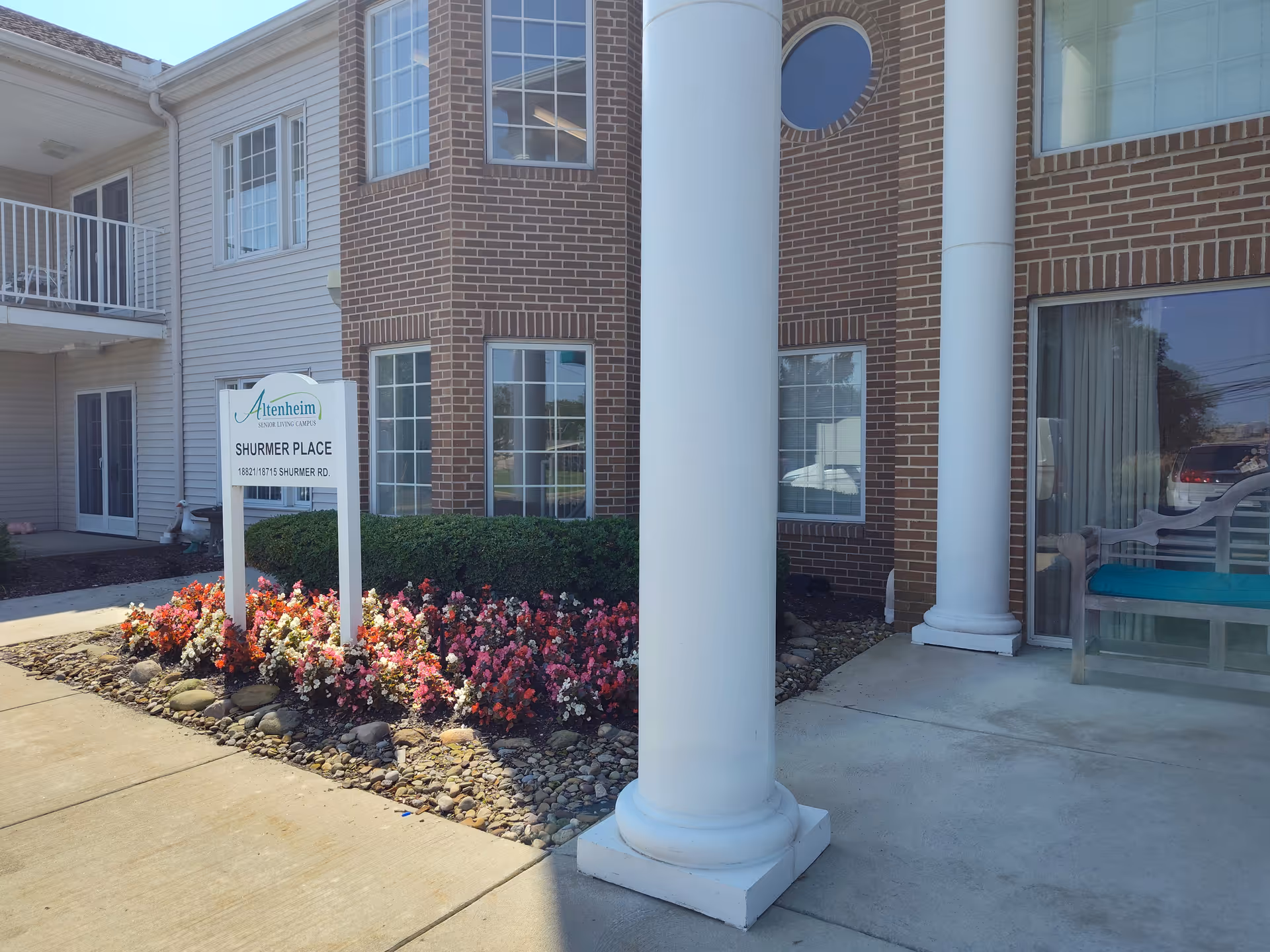 Exterior view of Shurmer Place at Altenheim with brick facade, columns, and flowering bed.