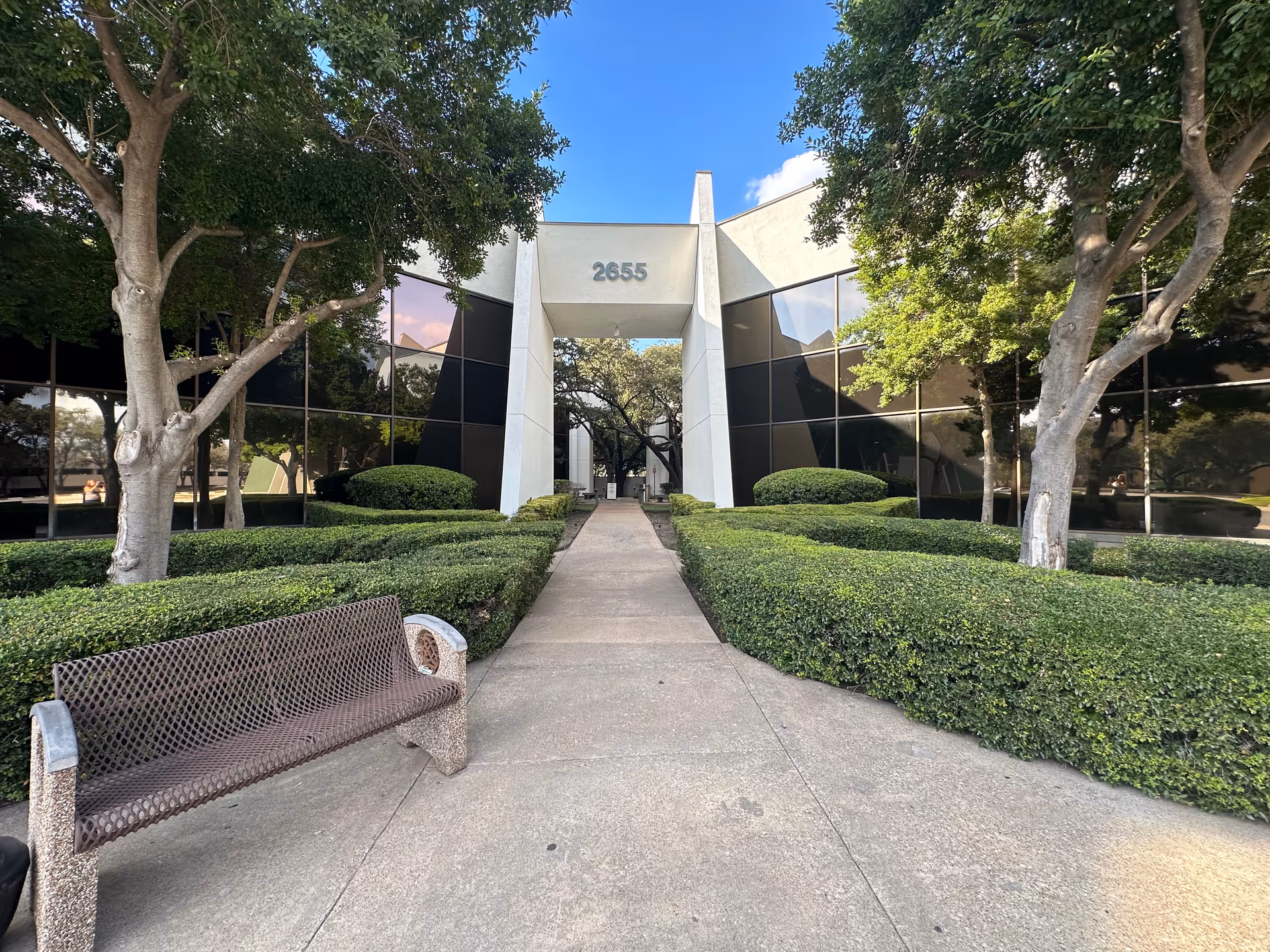 A wide concrete walkway leads to a modern building entrance, surrounded by hedges and trees.