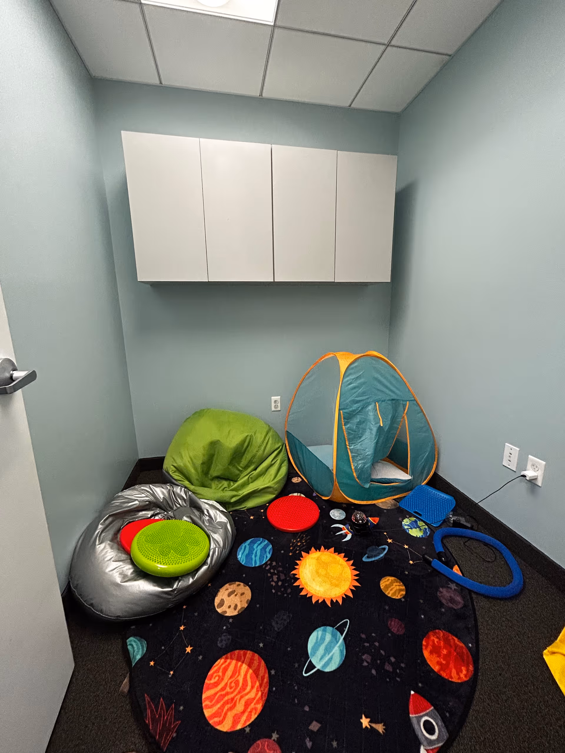 Small sensory room with space themed rug, bean bags, a blue tent, and wall cabinets.