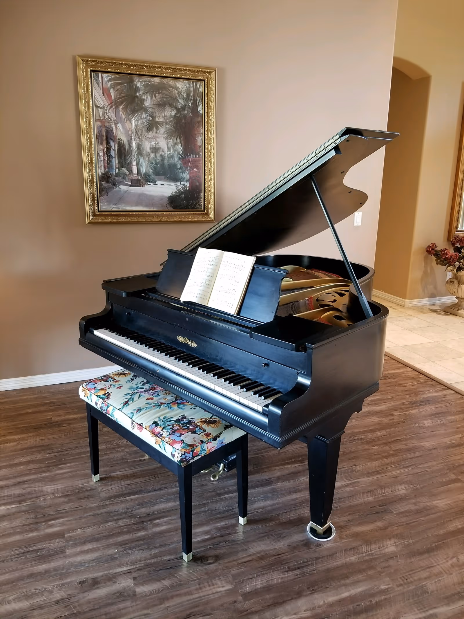 Black grand piano with its lid open and a floral upholstered bench on a wood floor beneath a framed painting.