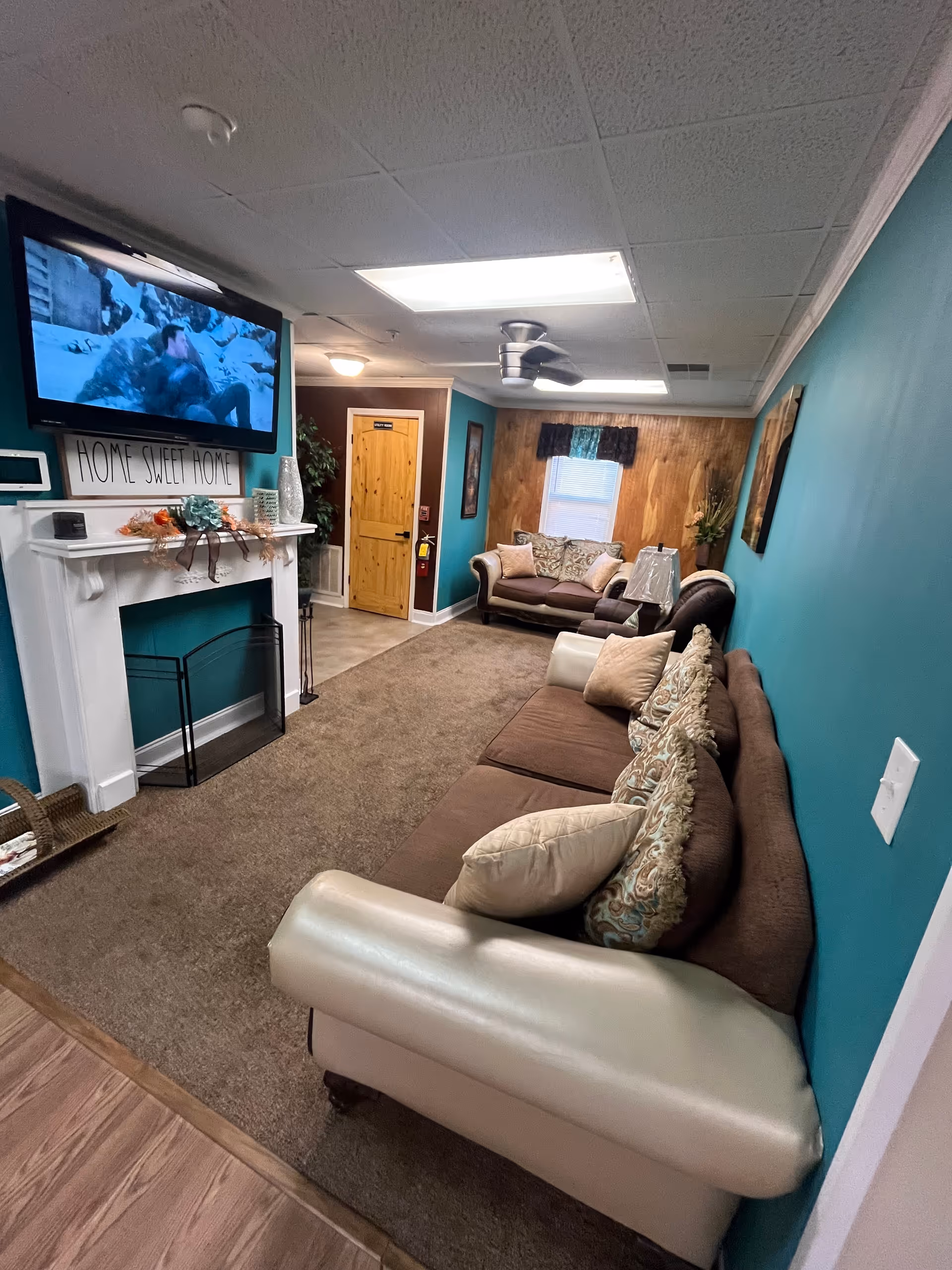 Cozy senior living lounge with teal walls, tan couches, fireplace, and mounted TV.