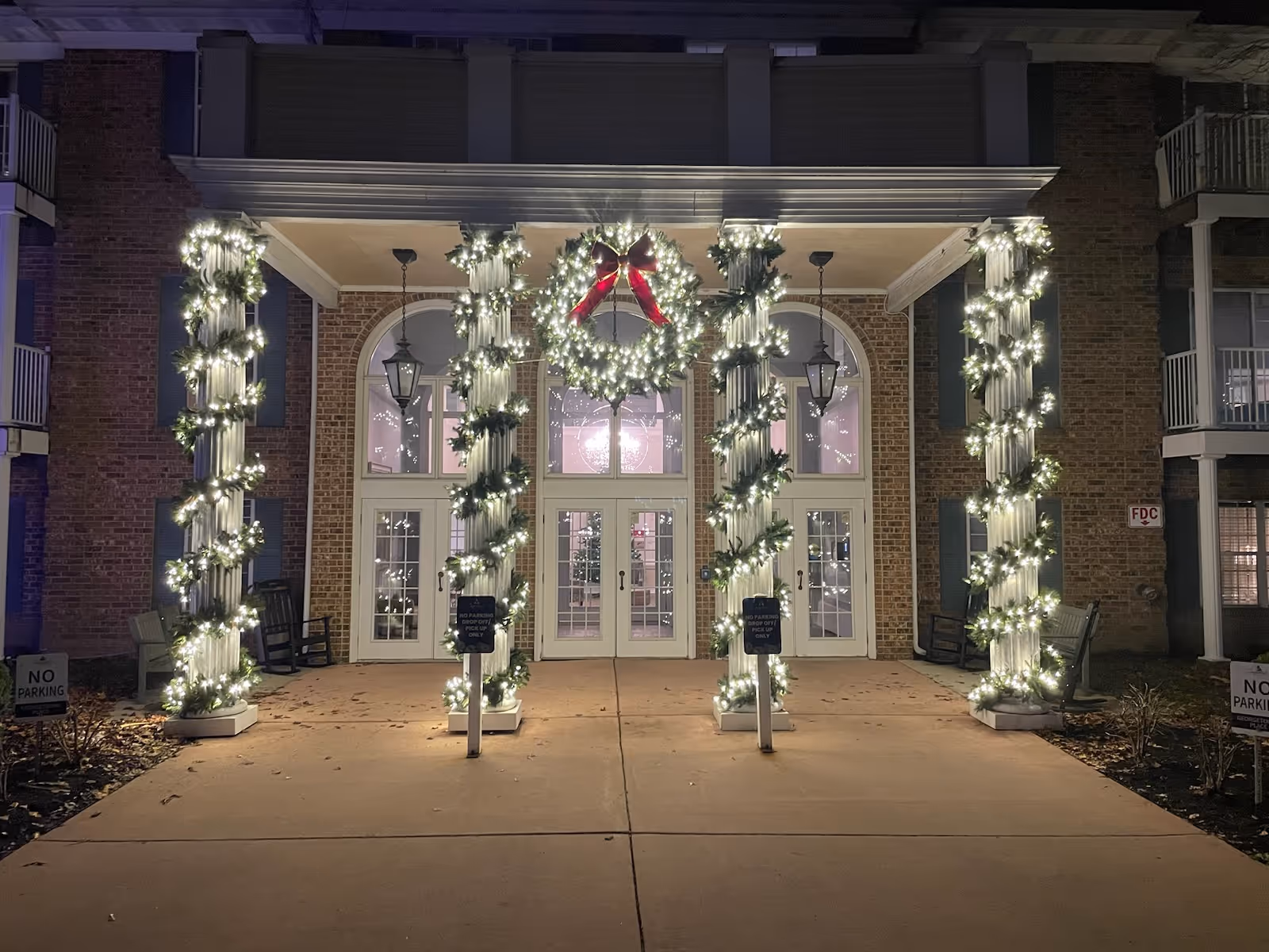 Nighttime exterior of a brick building entrance decorated with festively lit wrapped columns.