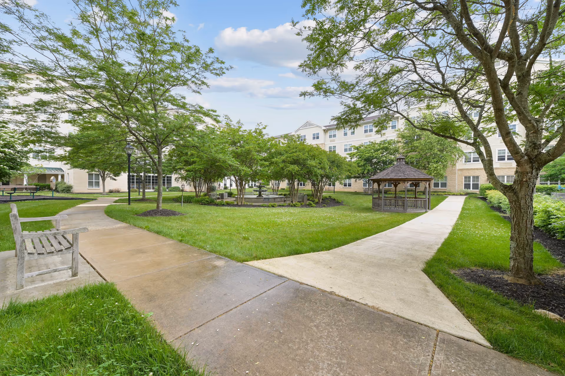 A neatly landscaped outdoor courtyard with paths, trees, a gazebo, and benches beside a building.