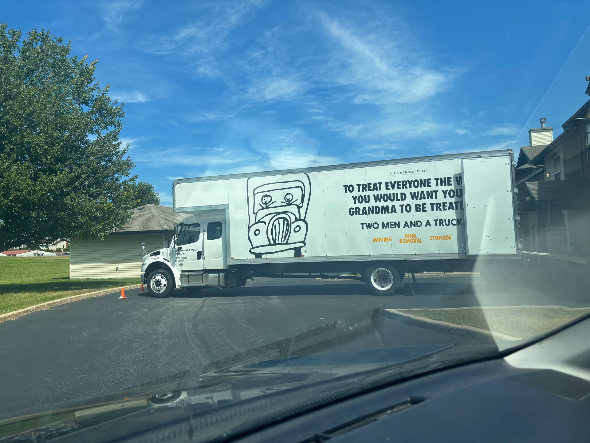White moving truck parked in a lot beside a green lawn and building.
