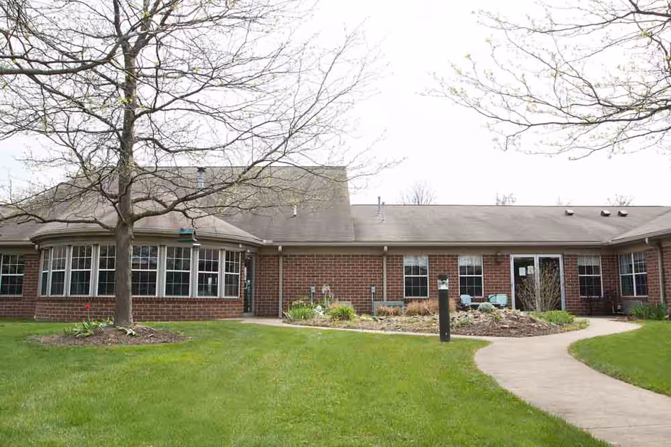 Exterior view of a single-story brick building with multiple windows and a sloped roof. There is a leafless tree in the foreground, a well-maintained green lawn, and a curved concrete walkway leading to the entrance. Some garden beds with plants and shrubs are visible near the building.