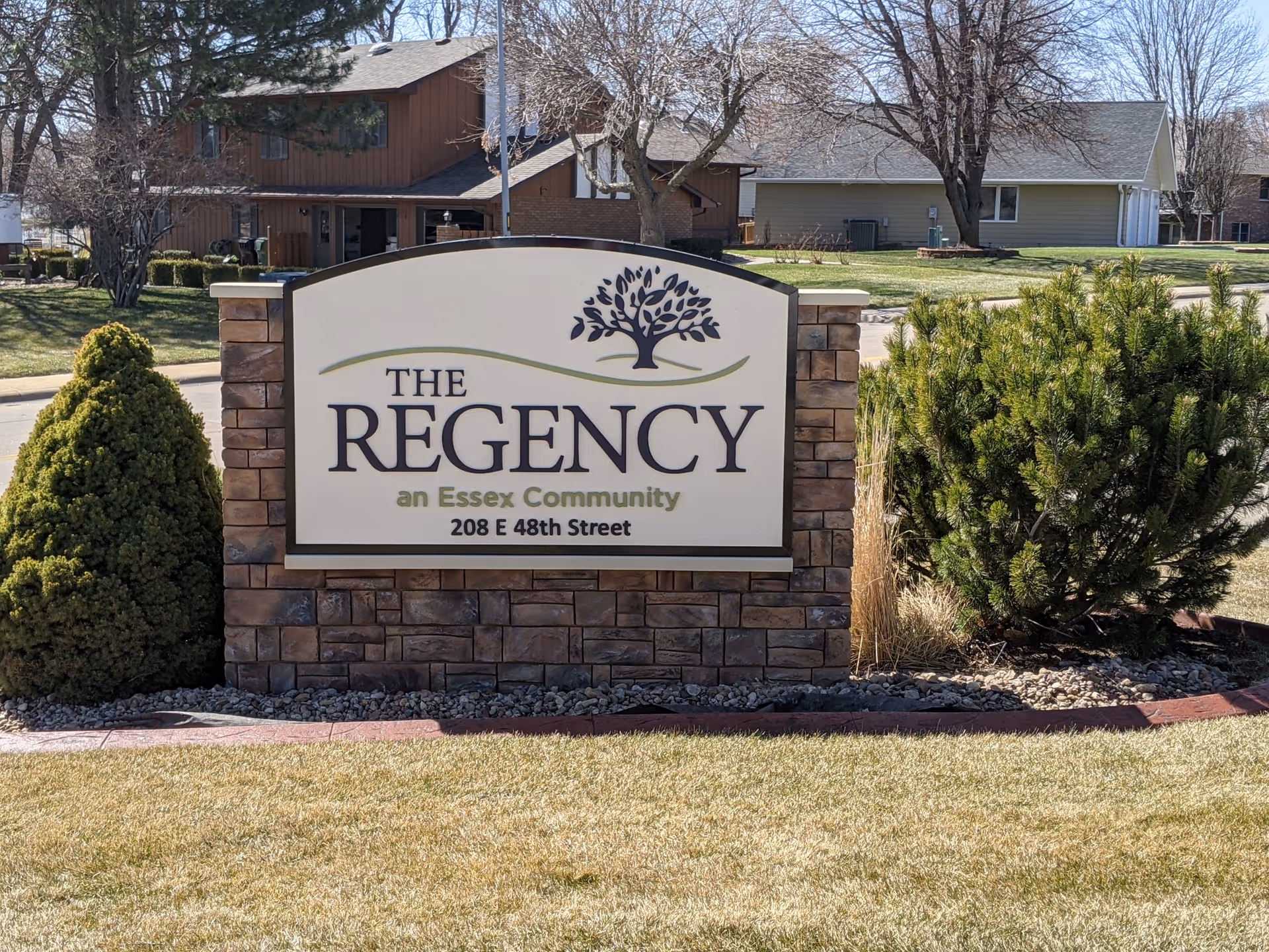 Outdoor sign reads The Regency, an Essex Community, with shrubs and houses in the background.